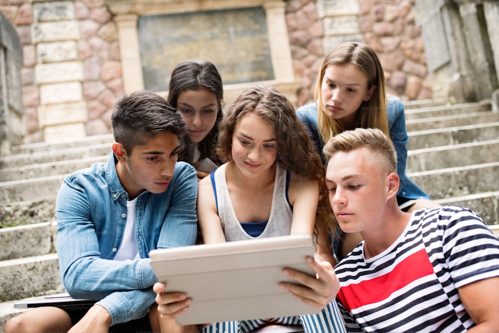 Group of attractive teenage students sitting on stone steps in front of university holding tablet, reading or watching something.