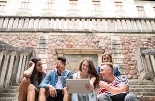 Group of attractive teenage students sitting on stone steps in front of university holding tablet, reading or watching something.
