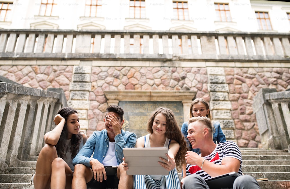 Group of attractive teenage students sitting on stone steps in front of university holding tablet, reading or watching something.