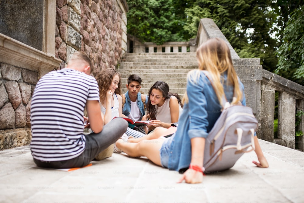 Groupe d’adolescents attrayants assis sur des marches de pierre devant l’université, lisant et étudiant.