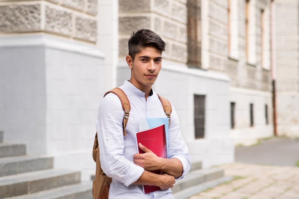 Handsome hispanic teenage student in white shirt standing in front of old building.