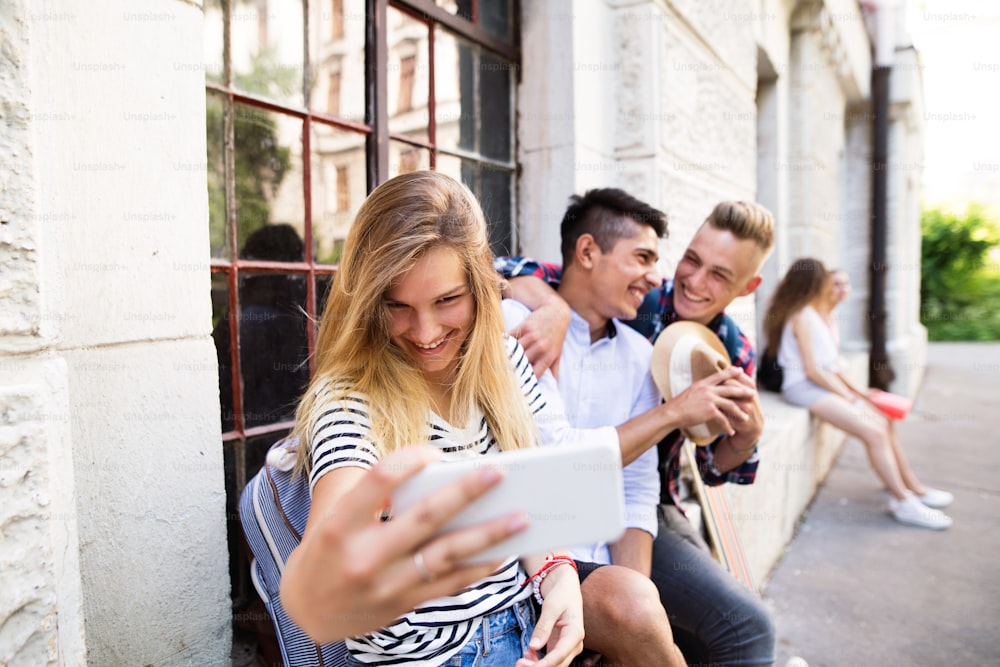 Group of attractive teenage students in front of university taking selfie with smart phone.