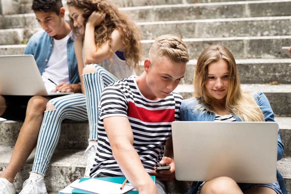 Attractive teenage students sitting on stone steps in front of university with laptops, reading or watching something.