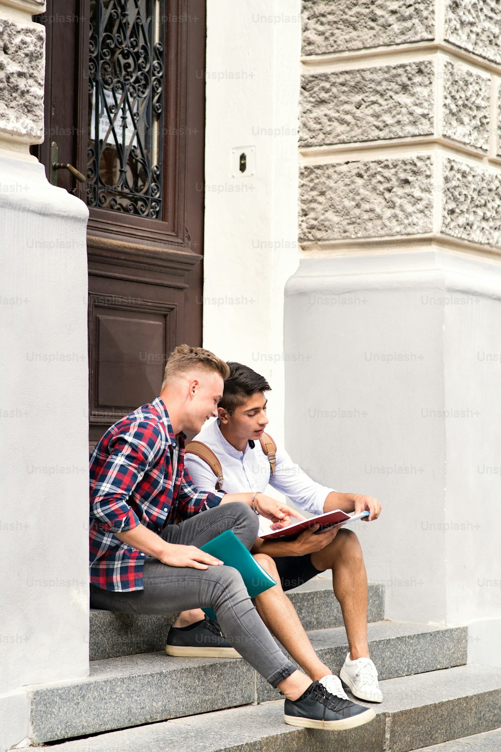 Two handsome teenage students sitting on stone steps in front of university.