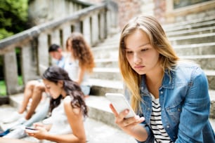 Atractiva estudiante adolescente sentada en escalones de piedra con sus amigos frente a la universidad sosteniendo un teléfono inteligente, leyendo o mirando algo.