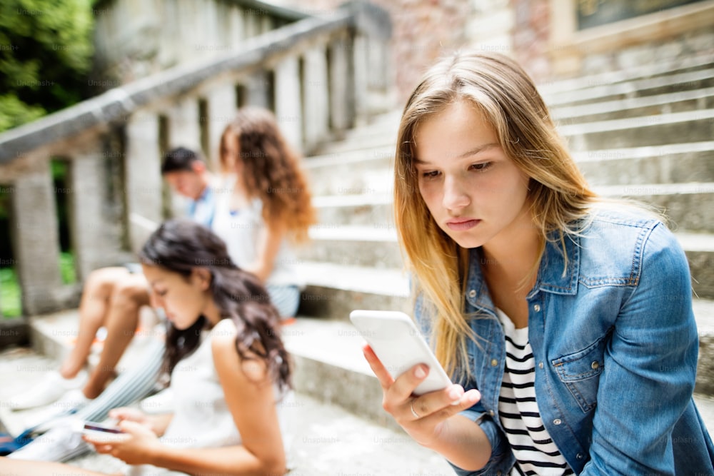 Attractive teenage student girl sitting on stone steps with her friends in front of university holding smart phone, reading or watching something.
