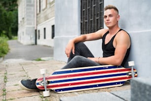 Cool teenage boy with scateboard sitting on stone path in front of old building.