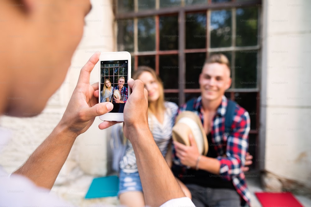 Unrecognizable man taking photo with smart phone of attractive teenage student couple posing in front of university.