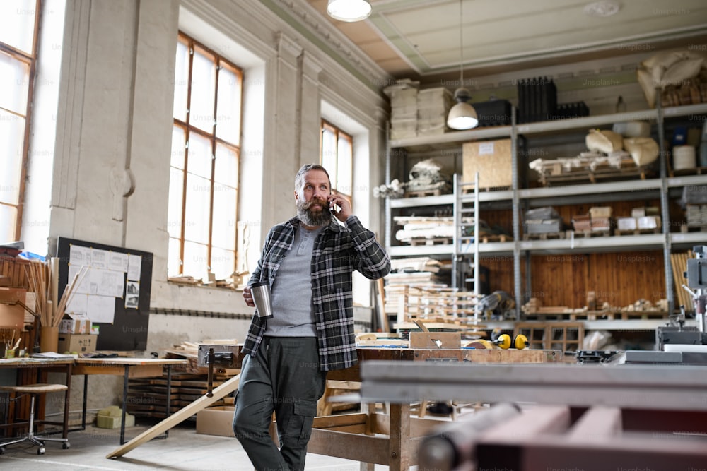 Un charpentier mûr heureux faisant un appel téléphonique pendant la pause-café à l’intérieur dans un atelier de menuiserie.