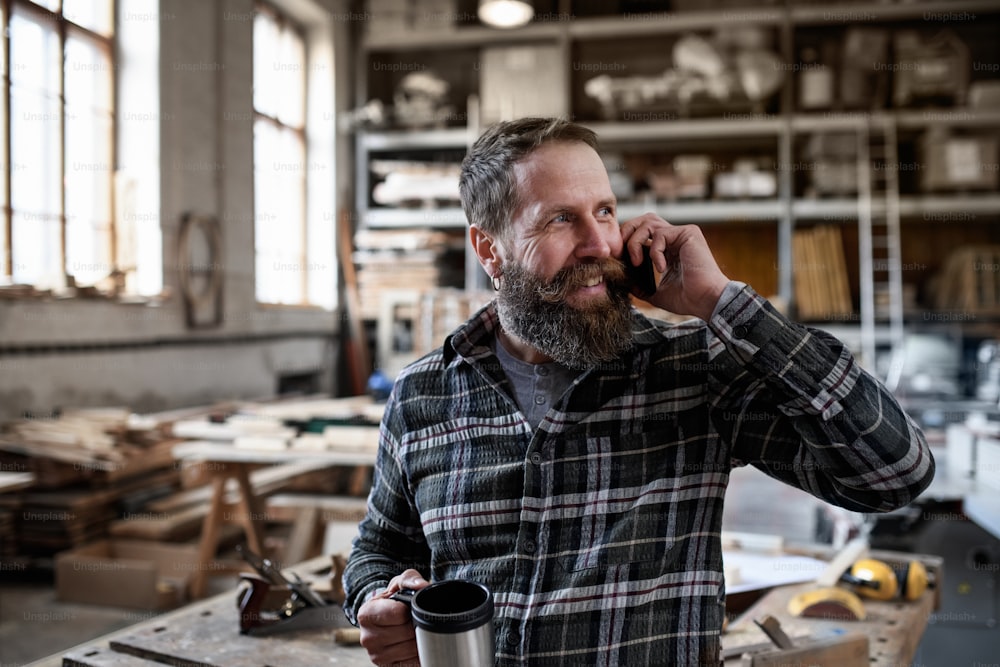 Um carpinteiro maduro e feliz fazendo telefonema durante a pausa para o café dentro de casa na oficina de carpentery.