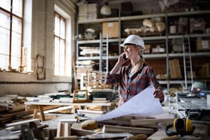 Una ingeniera sosteniendo planos y haciendo llamadas telefónicas en el interior de un taller de carpintería.