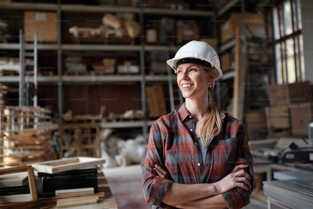 A portrait of midadult female carpenter standing in carpentery workshop, looking aside and smiling. Small business concept.