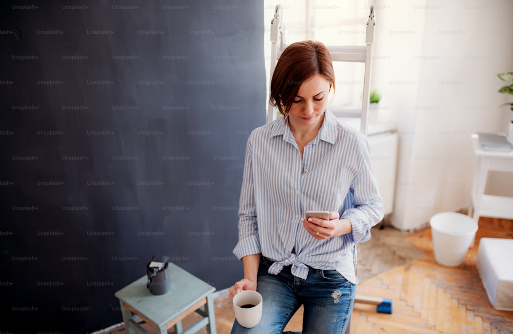Un retrato de una joven mujer creativa con una taza de café y un teléfono inteligente pintando la pared de negro. Una startup de pequeñas empresas.