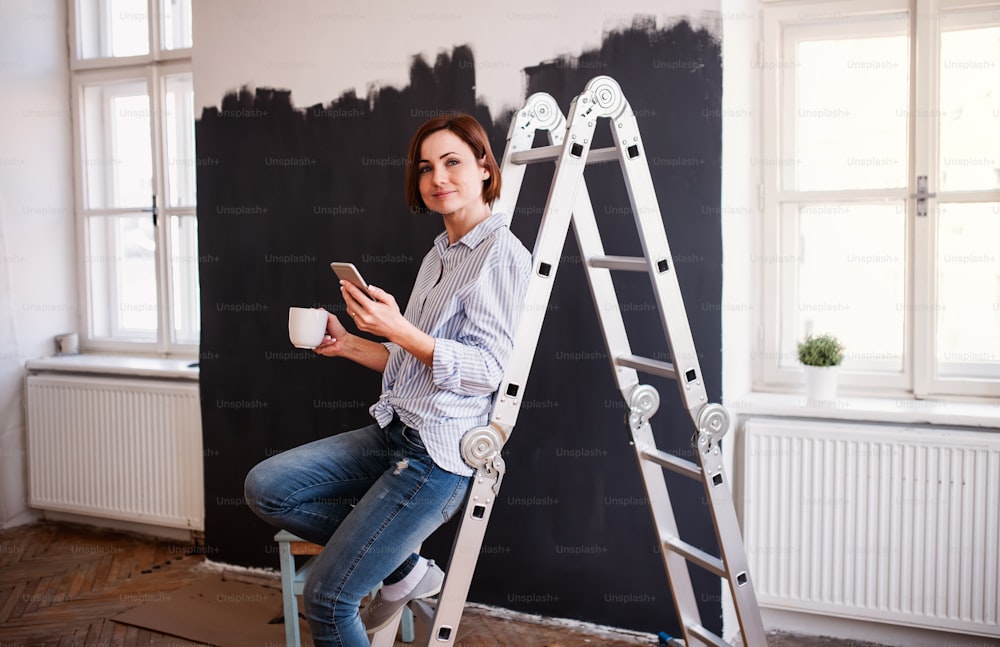 Um retrato de uma jovem mulher criativa com uma xícara de café e smartphone pintando parede preta. Uma startup de pequenas empresas.