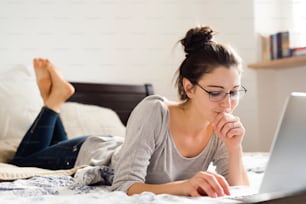 Beautiful young woman lying on bed, working on notebook, home office.