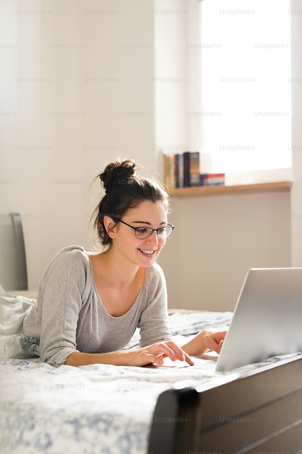 안경을 쓴 아름다운 젊은 여자가 침대에 누워 노트북과 홈 오피스에서 일하고 있다.