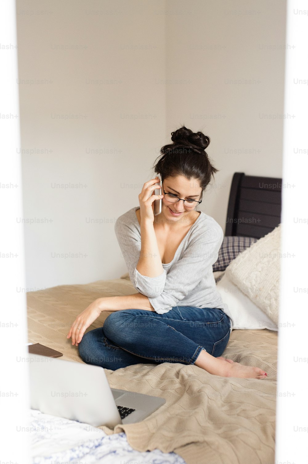 Bella giovane donna che indossa occhiali da vista seduta sul letto, taccuino accanto a lei, tenendo in mano lo smartphone, facendo una telefonata, ufficio a casa.