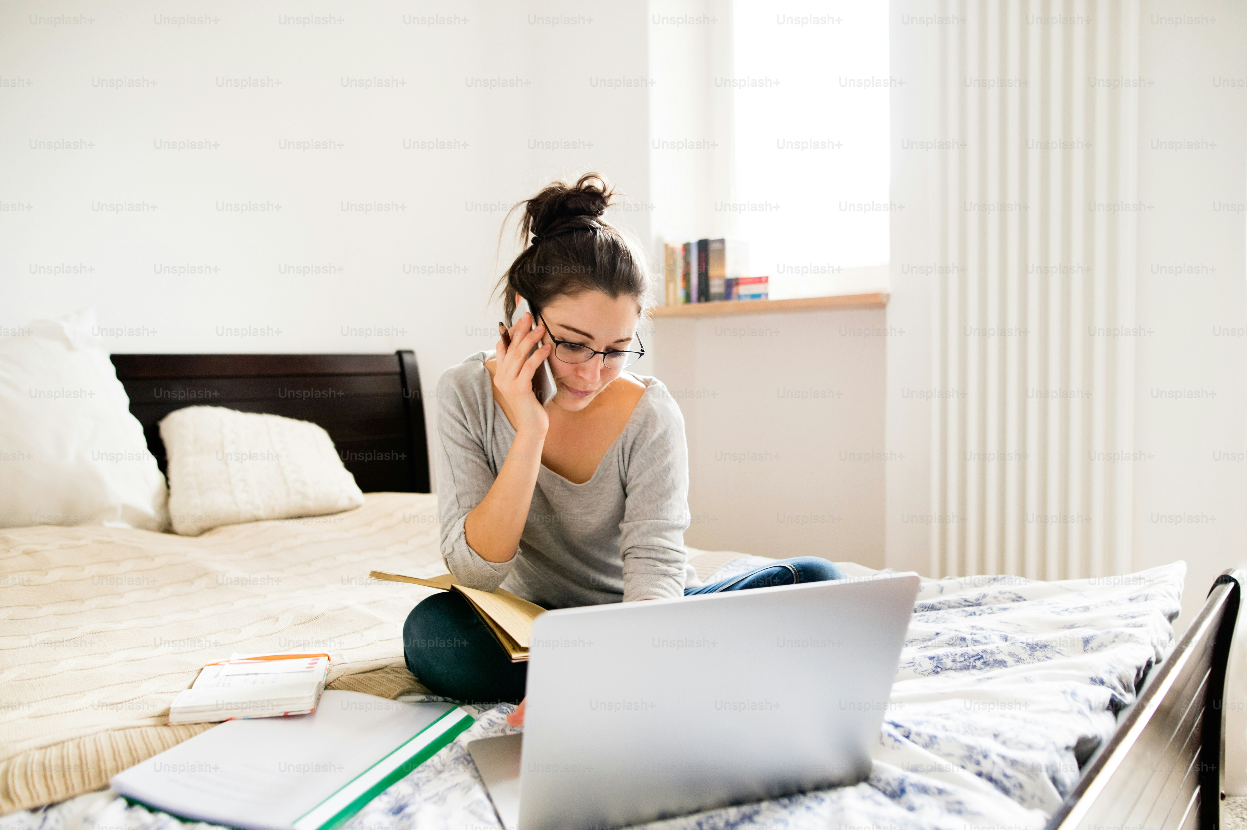 Wanita muda cantik mengenakan kacamata duduk di tempat tidur, mengerjakan notebook, memegang ponsel pintar, melakukan panggilan telepon, kantor rumah.