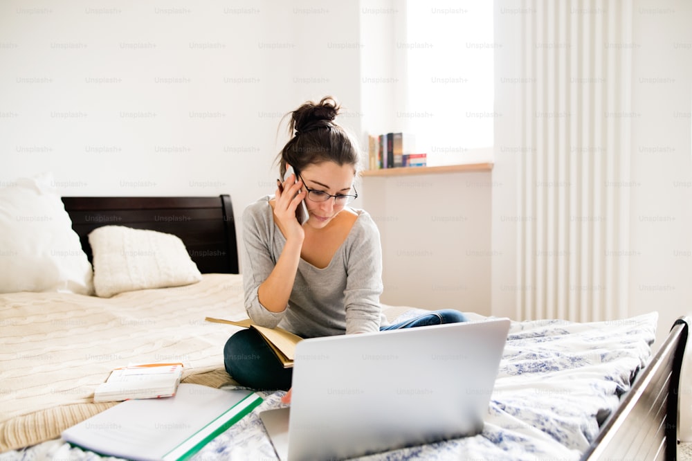 Schöne junge Frau mit Brille, die auf dem Bett sitzt, an einem Notebook arbeitet, ein Smartphone hält, telefoniert, Home-Office.