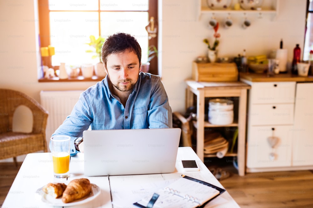 Hombre sentado en la mesa de la cocina trabajando desde casa en una computadora portátil