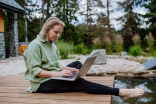 노트북으로 일하는 프리랜서인 한 여성이 뒤뜰 호수 옆 부두에 앉아 원격 사무실의 개념으로 휴가 중에 일합니다.