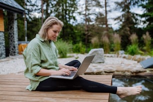 A woman, freelancer working on laptop, sitting on a pier by the backyard lake, a concept of remote office, work during vacation.