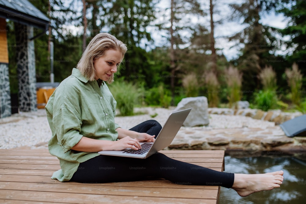 A woman, freelancer working on laptop, sitting on a pier by the backyard lake, a concept of remote office, work during vacation.