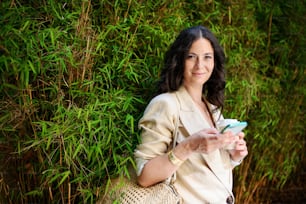 A happy woman traveller, texting on mobile phone and holding cofee cup, with green plant at background.