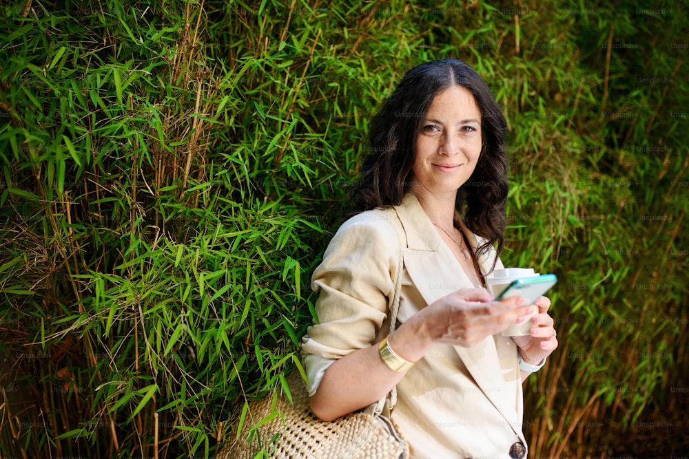 Eine glückliche Reisende, die eine SMS auf dem Handy schreibt und einen Cofee-Becher in der Hand hält, mit grüner Pflanze im Hintergrund.