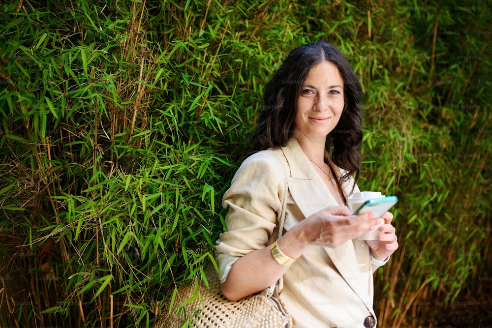 행복한 여성 여행자, 휴대 전화로 문자 메시지를 보내고 코피 컵을 들고 배경에 녹색 식물이 있습니다.