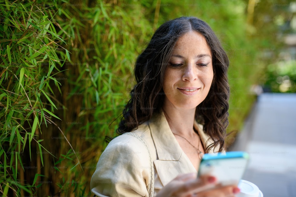 Uma mulher feliz viajante, enviando mensagens de texto no celular e segurando copo de cofee, com planta verde ao fundo.