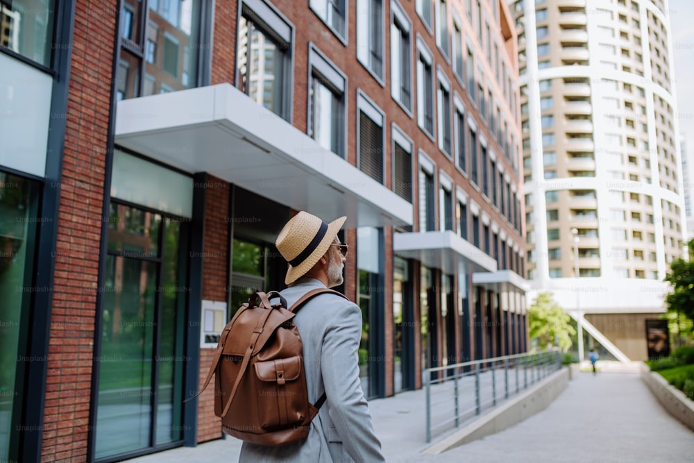 Uma visão traseira de homem usando chapéu de palha e mochila andando na rua, empresário em roupas casuais no verão a caminho do trabalho.