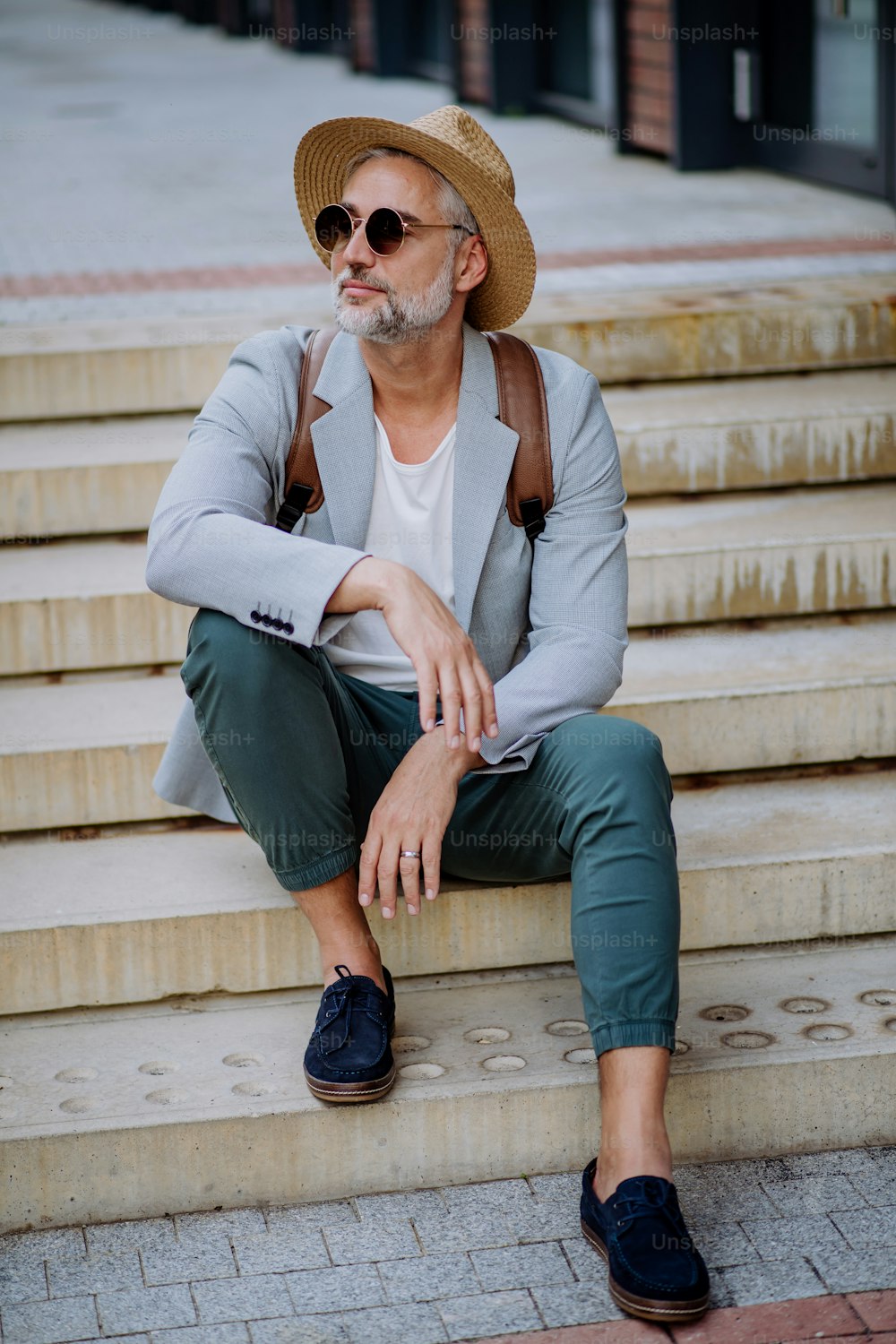밀짚모자와 배낭을 메고 계단에 앉아 있는 자신감 넘치는 남자, 여름에 캐주얼한 옷을 입고 출근하는 사업가.