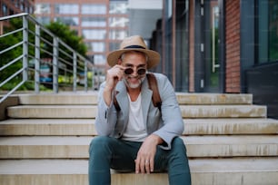 Un portrait d’un homme confiant portant un chapeau de paille et un sac à dos assis dans les escaliers et regardant la caméra, homme d’affaires en tenue décontractée en été sur le chemin du travail.