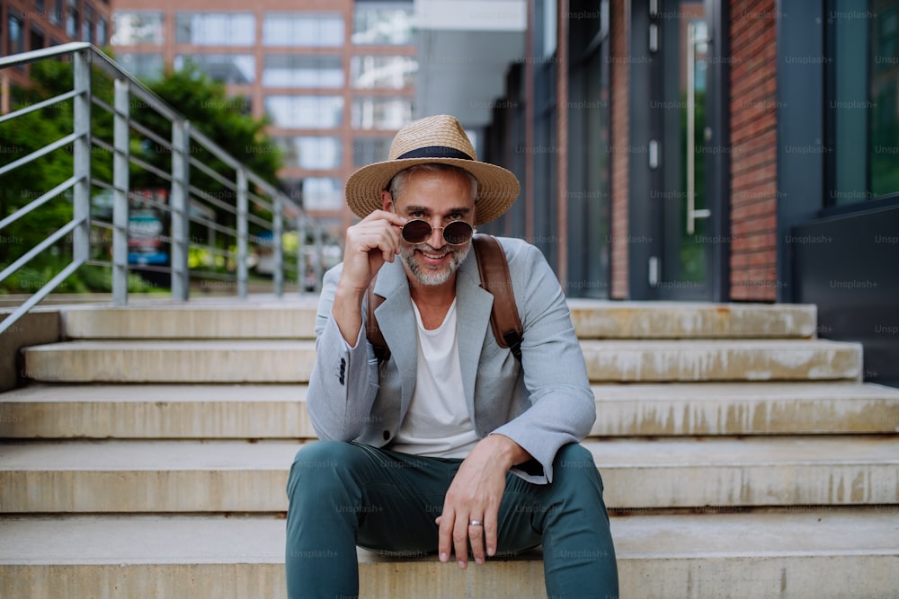 A portrait of confident man wearing straw hat and backpack sitting on stairs and looking at camera, businessman in casual clothes in summer on the way to work.