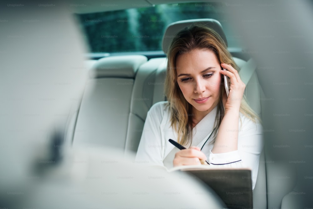 Mujer de negocios con teléfono inteligente sentada en los asientos traseros en un taxi, haciendo una llamada telefónica.