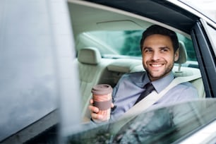 Young businessman with coffee sitting on back seat in car.