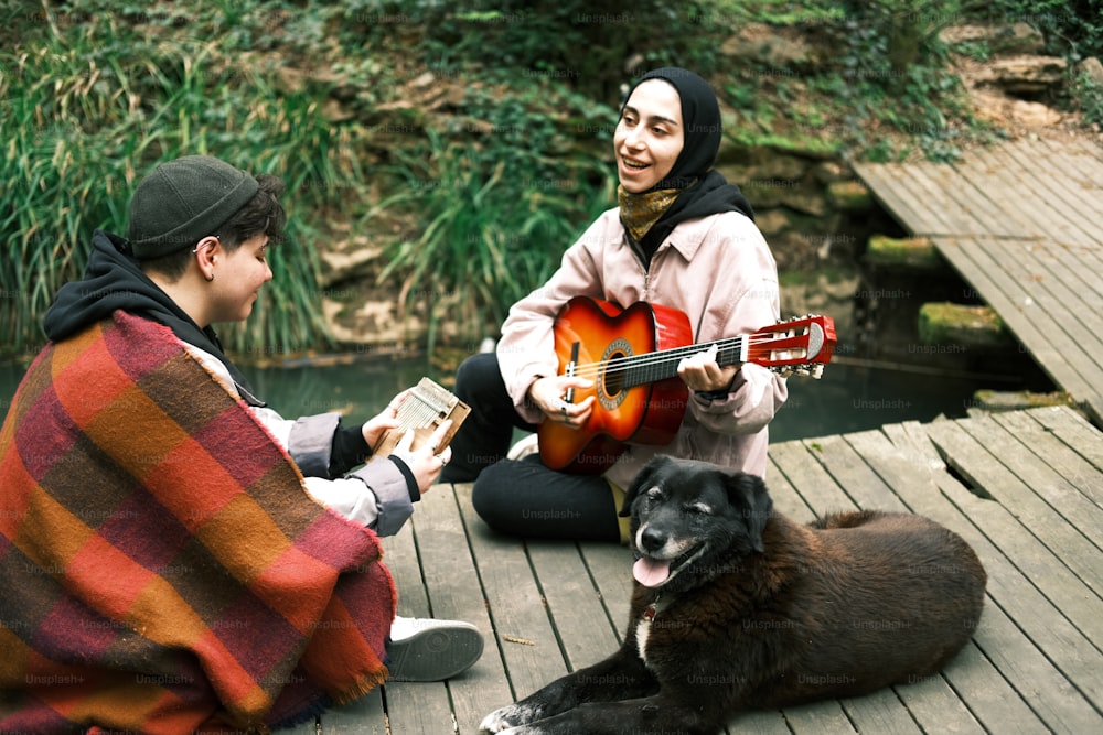 two people sitting on a dock with a dog and a guitar