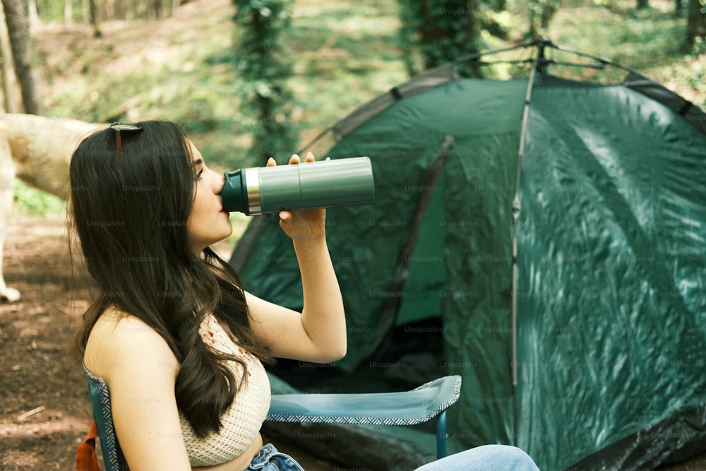 a woman sitting in front of a tent drinking from a cup