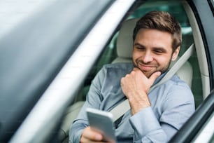 Joven hombre de negocios con camisa, corbata y teléfono inteligente sentado en el automóvil, mensajes de texto.