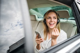 Mujer de negocios con teléfono inteligente y auriculares sentada en los asientos traseros en un taxi, escuchando música.