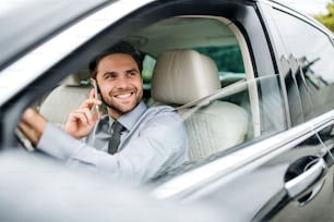 Joven empresario con camisa, corbata y teléfono inteligente sentado en el automóvil, haciendo una llamada telefónica.