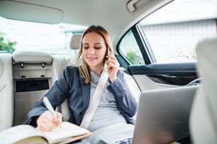 Mujer de negocios con teléfono inteligente y computadora portátil sentada en los asientos traseros en un taxi, hablando por teléfono.