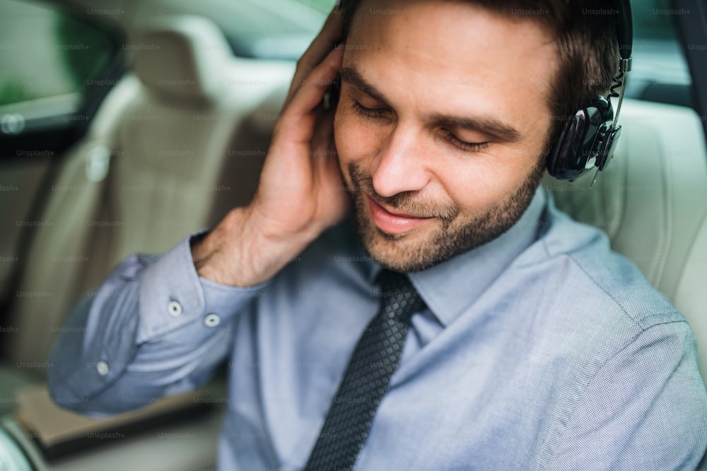 Giovane uomo d'affari felice con cuffie, camicia e cravatta seduto in auto, ascoltando musica.