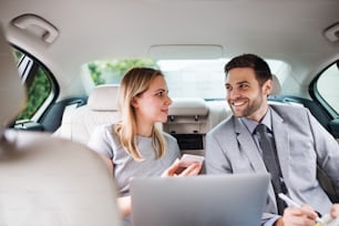 Young business couple with laptop sitting on back seats in car, talking.