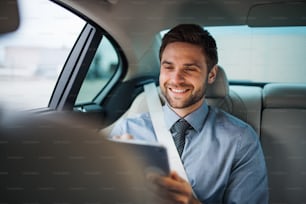 A businessman with tablet sitting on back seats in car, working.
