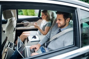 Young business couple with laptop sitting on back seats in car, working.