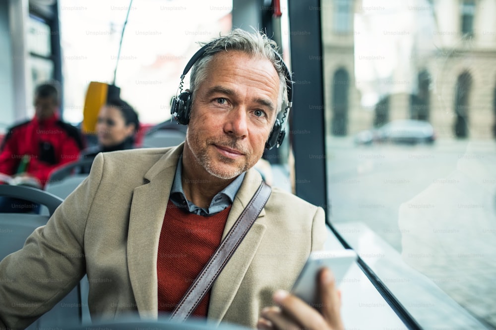 Hombre de negocios maduro y cansado con teléfono inteligente y heaphones viajando en autobús en la ciudad, escuchando música.
