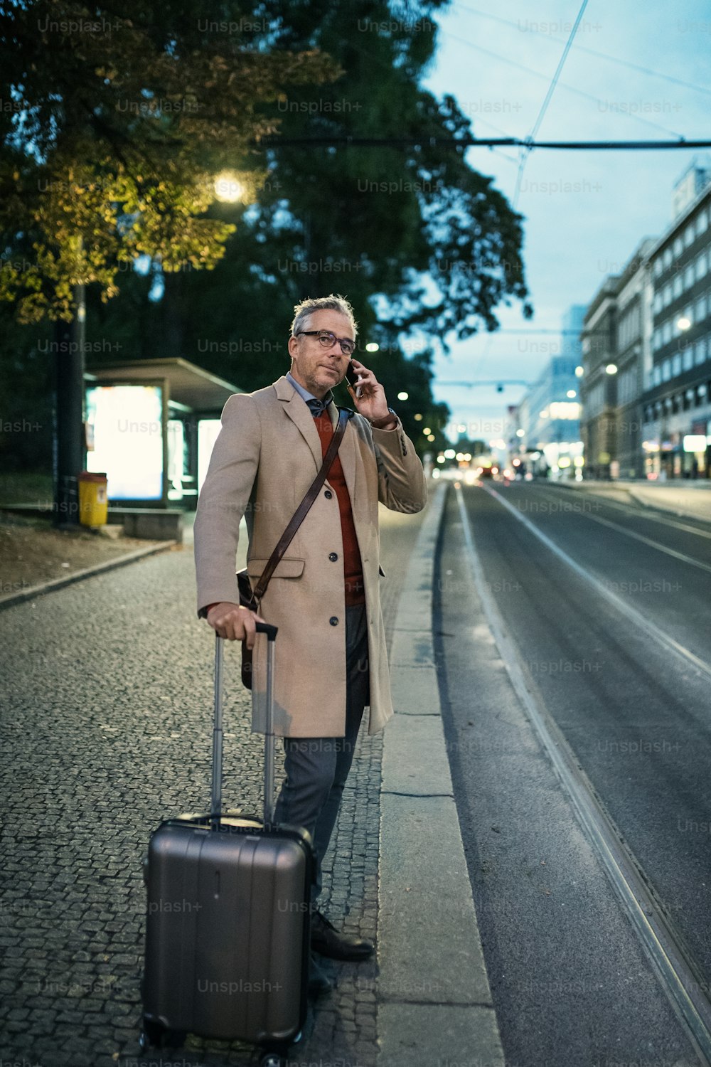 スマートフォンとスーツケースを持つ成熟した実業家が、プラハ市で夕方に路面電車やトロリー車を待っていて、電話をかける。