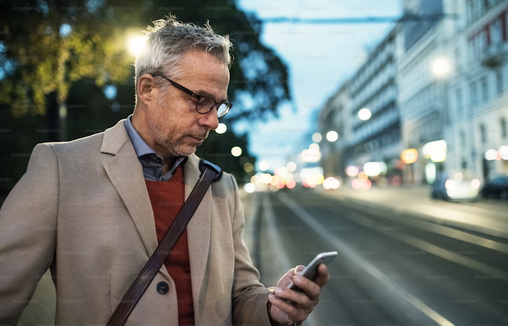 Un uomo d'affari maturo con uno smartphone in piedi su una strada la sera, che manda messaggi. Copia spazio.